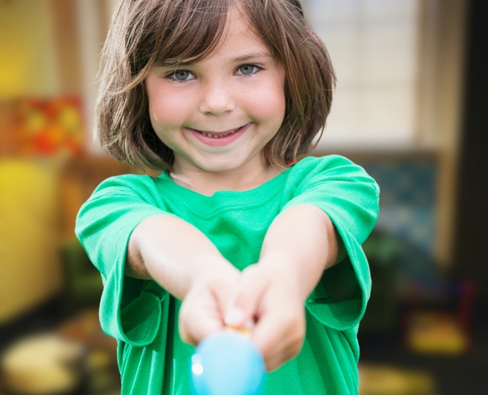 A young child with brown hair and a green shirt smiles while holding a blue object toward the camera in a brightly lit indoor setting.